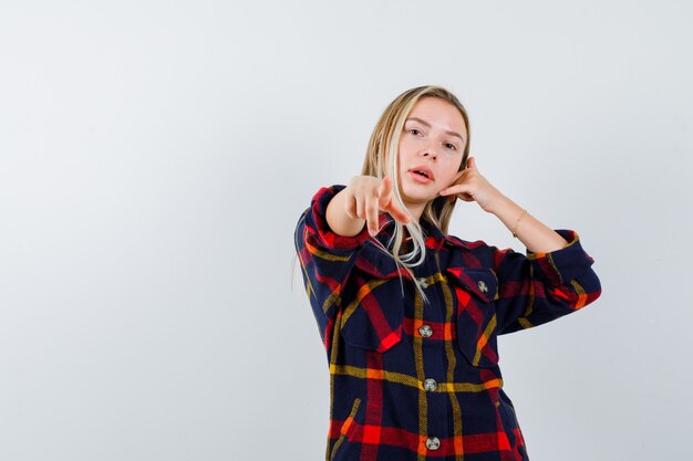 Jeune femme en chemise à carreaux pointant la caméra tout en tenant la main sur la tête et à la vue de face, confiant.