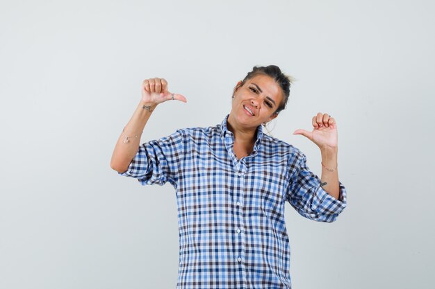 Jeune femme en chemise à carreaux pointant sur elle-même et à la joyeuse.