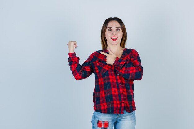 Jeune femme en chemise à carreaux pointant vers l'arrière et à la surprise, vue de face.