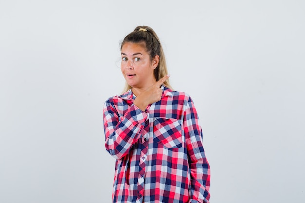 Jeune femme en chemise à carreaux pointant vers le coin supérieur droit et à la vue de face, hésitante.