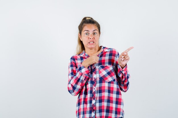Jeune femme en chemise à carreaux pointant vers le côté droit et regardant troublé, vue de face.