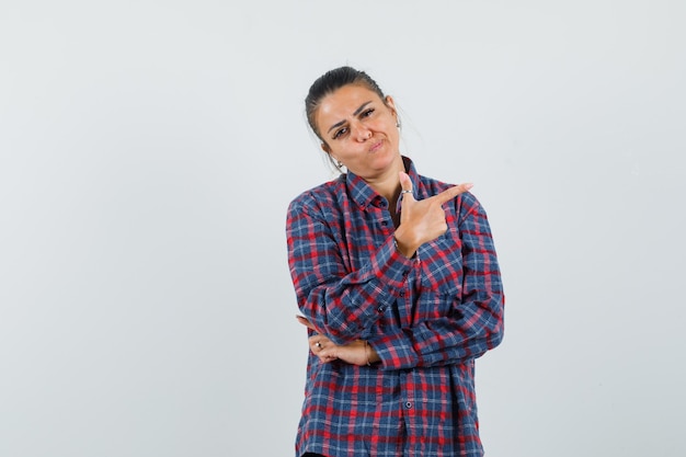 Jeune femme en chemise à carreaux pointant vers la droite avec l'index et regardant pensif, vue de face.