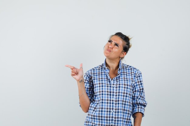 Jeune femme en chemise à carreaux pointant vers l'extérieur et à la pensif.