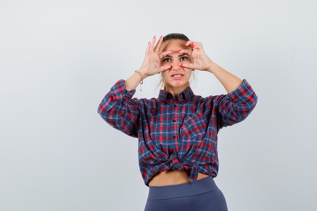 Jeune femme en chemise à carreaux regardant à travers les doigts et l'air curieux , vue de face.