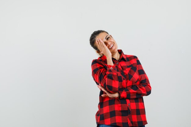 Jeune femme en chemise à carreaux tenant la main sur le visage en riant, vue de face.