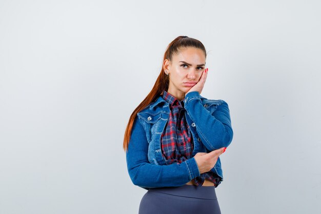 Jeune femme en chemise à carreaux, veste en jean appuyée sur la joue et l'air ennuyée, vue de face.