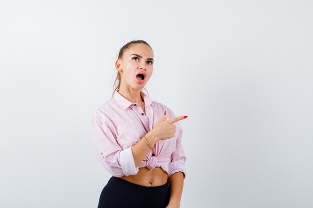 Jeune femme en chemise décontractée pointant vers le côté droit et regardant perplexe, vue de face.