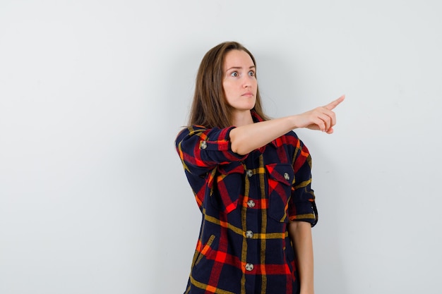 Jeune femme en chemise décontractée pointant vers l'extérieur et regardant focalisée, vue de face.