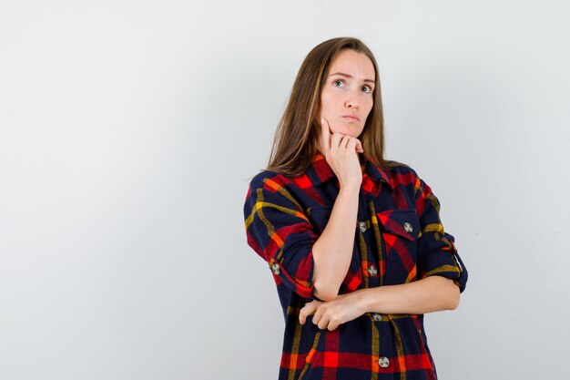 Jeune femme en chemise décontractée soutenant le menton sur place et regardant pensif, vue de face.