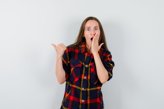 Jeune femme en chemise décontractée tenant la main sur la bouche ouverte, pointant de côté et regardant choqué, vue de face.