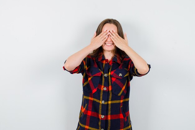 Jeune femme en chemise décontractée, tenant les mains sur les yeux et à la vue de face, joyeuse.