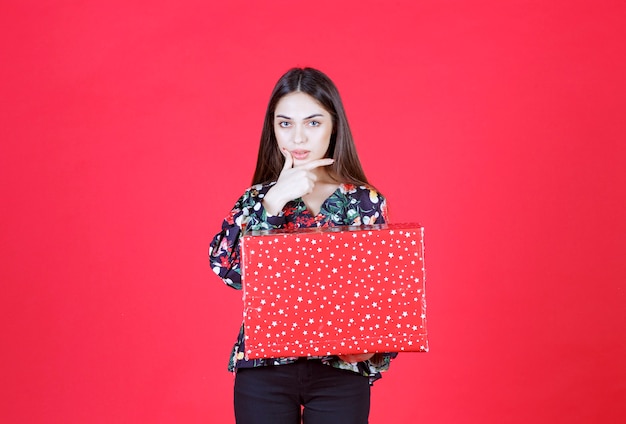 Jeune femme en chemise à fleurs tenant une boîte-cadeau rouge avec des points blancs dessus et a l'air confuse et réfléchie