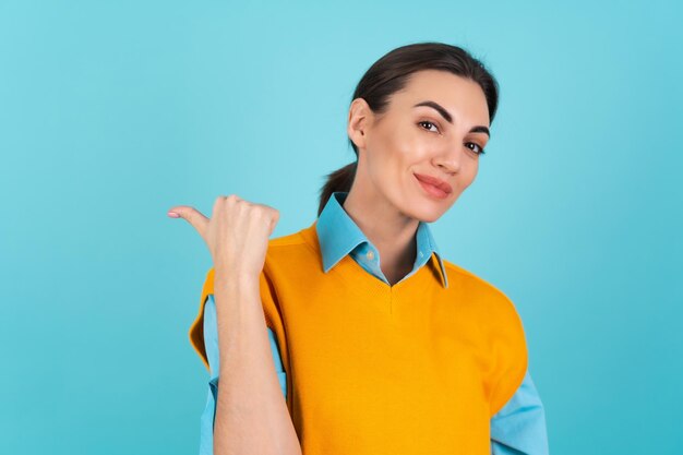 Jeune femme en chemise et gilet orange sur fond turquoise souriant joyeusement, pointant son doigt vers la gauche vers un espace vide