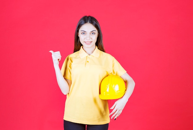 Jeune femme en chemise jaune tenant un casque jaune et montrant quelqu'un derrière