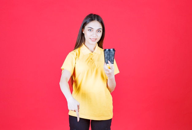 Jeune femme en chemise jaune tenant une tasse de café jetable noire et invitant son partenaire à partager avec