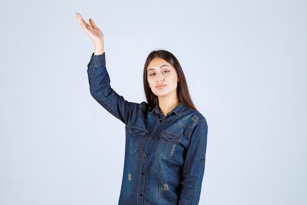 Jeune femme en chemise en jean levant les mains et montrant quelque chose au-dessus