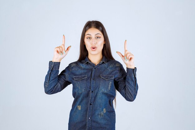 Jeune femme en chemise en jean levant les mains et montrant quelque chose au-dessus