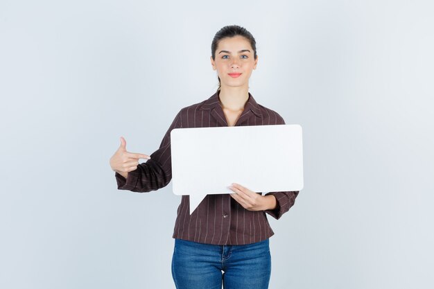 Jeune femme en chemise, jeans pointant sur le côté, gardant une affiche en papier et ayant l'air satisfaite, vue de face.