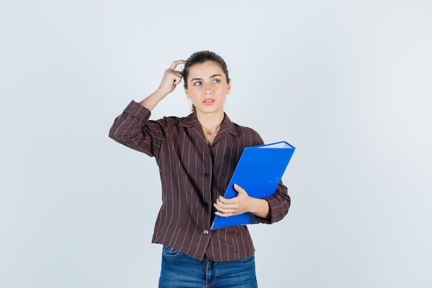 Jeune femme en chemise, jeans tenant un dossier, debout dans une pose de réflexion et semblant raisonnable, vue de face.