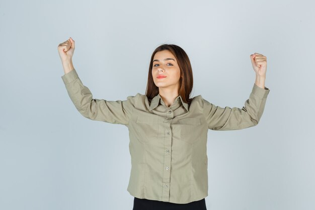 Jeune femme en chemise, jupe montrant le geste du gagnant et semblant chanceuse, vue de face.