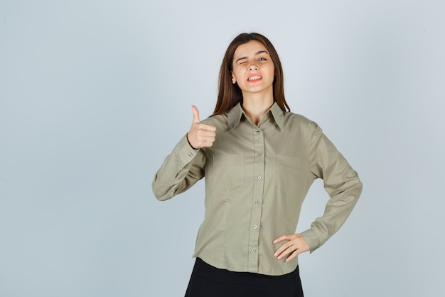 Jeune femme en chemise, jupe montrant le pouce vers le haut tout en faisant un clin d'œil et l'air heureux, vue de face.