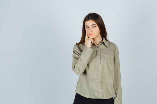 Jeune femme en chemise, jupe pointant sur sa paupière et ayant l'air fatiguée, vue de face.