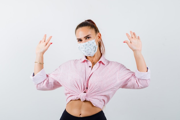 Jeune femme en chemise, pantalon, masque médical montrant les paumes en geste de reddition et à la vue de face sérieuse.