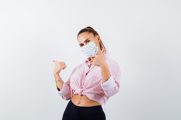 Jeune femme en chemise, pantalon, masque montrant le double pouce vers le haut et l'air heureux, vue de face.