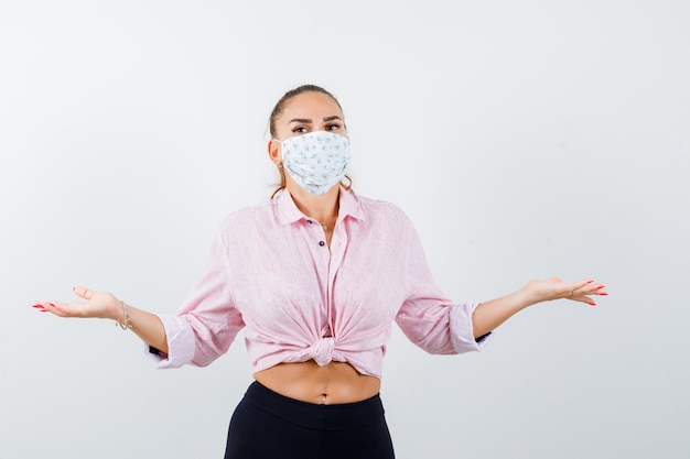 Jeune femme en chemise, pantalon, masque montrant un geste impuissant et regardant perplexe, vue de face.