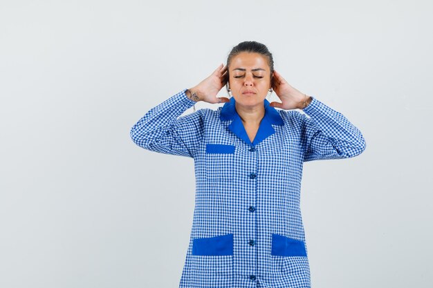 Jeune femme en chemise de pyjama vichy bleu levant les mains près de la tête tout en fermant les yeux et à la vue détendue, de face.