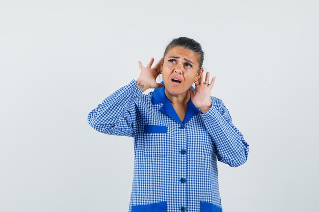 Jeune femme en chemise de pyjama vichy bleu main dans la main près des oreilles et à la recherche de stress, vue de face.