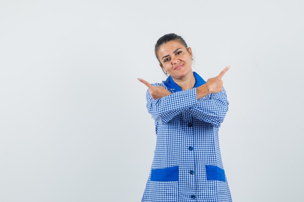 Photo gratuite jeune femme en chemise de pyjama vichy bleu pointant dans des directions opposées avec l'index et à la jolie vue de face.