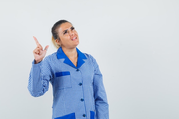 Jeune femme en chemise de pyjama vichy bleu pointant vers le haut et regardant pensif, vue de face.