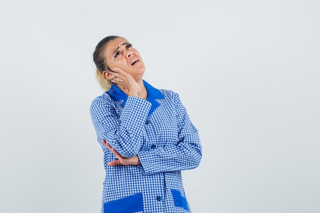 Jeune femme en chemise de pyjama vichy bleu se penchant la main sur la joue, debout dans la pensée pose et à la vue épuisée, de face.