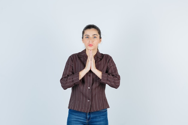 Photo gratuite jeune femme en chemise rayée, jeans montrant le geste de namaste et l'air sérieux, vue de face.