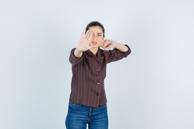 Jeune femme en chemise rayée, jeans montrant un panneau d'arrêt, serrant le poing et regardant en colère, vue de face.