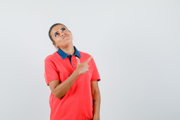 Jeune femme en chemise rouge pointant le côté supérieur droit avec l'index et regardant au-dessus et à la jolie vue de face.