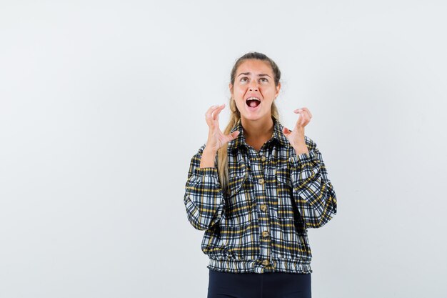 Jeune femme en chemise, short levant les mains de manière agressive tout en criant, vue de face.