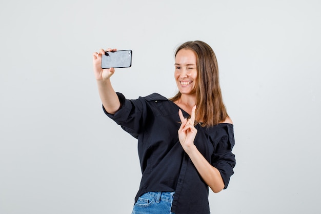 Jeune femme en chemise, short montrant v-sign tout en prenant selfie et à la joie