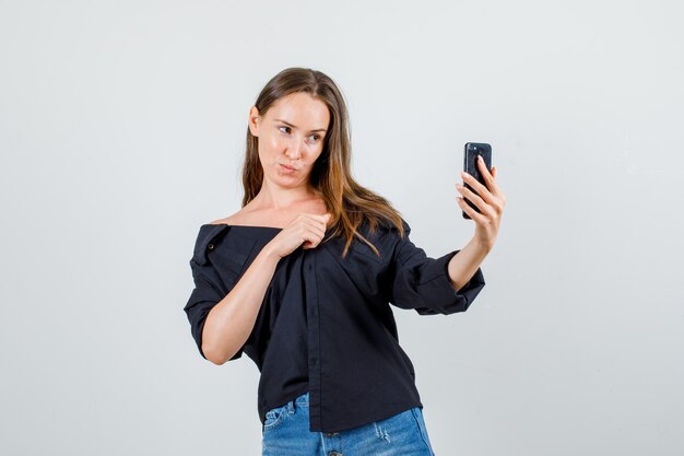 Jeune femme en chemise, short posant tout en prenant selfie sur smartphone et à mignon