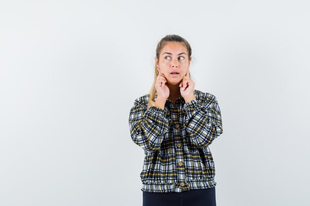 Jeune femme en chemise, short tenant les doigts sur les joues et à la rêveuse, vue de face.