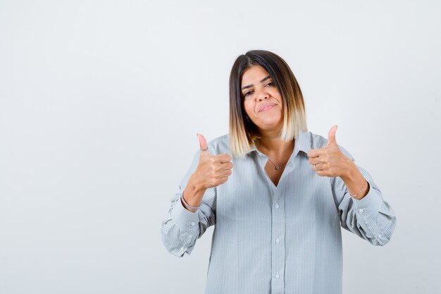 Jeune femme en chemise surdimensionnée montrant les pouces vers le haut et l'air heureux, vue de face.