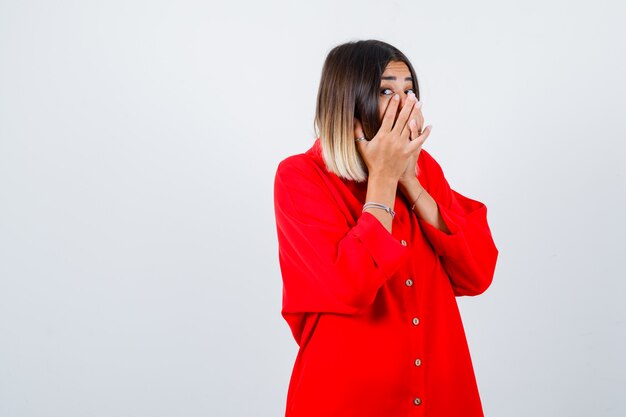 Jeune femme en chemise surdimensionnée rouge avec les mains sur la bouche et l'air perplexe, vue de face.