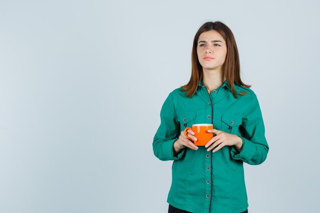 Jeune femme en chemise tenant une tasse de thé orange et regardant focalisée, vue de face.
