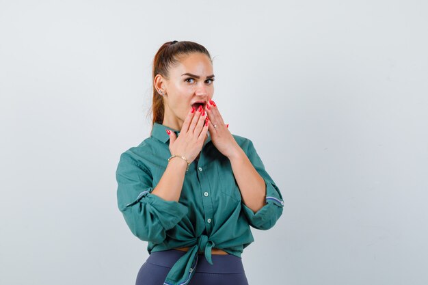 Jeune femme en chemise verte avec les mains sur la bouche ouverte et l'air surpris, vue de face.