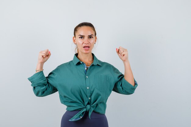 Jeune femme en chemise verte montrant le geste du gagnant et ayant l'air chanceux, vue de face.