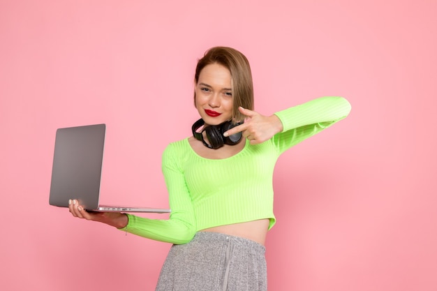 jeune femme en chemise verte et pantalon gris posant avec ordinateur portable et écouteurs noirs