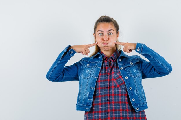 Photo gratuite jeune femme en chemise, veste en appuyant sur les doigts sur les joues soufflées et à la drôle, vue de face.