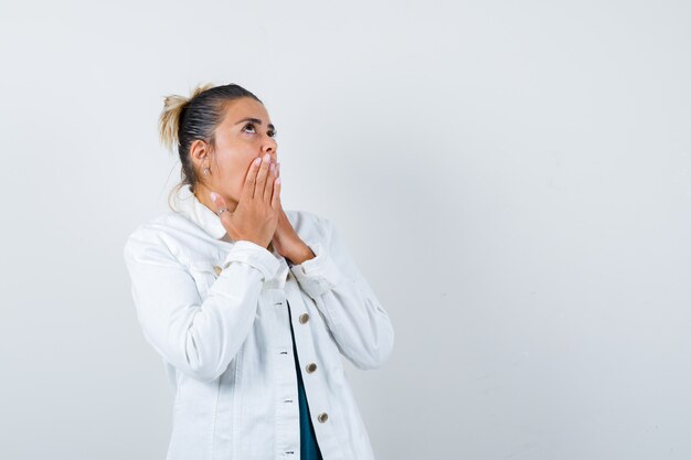 Jeune femme en chemise, veste blanche avec les mains sur la bouche et l'air pensif, vue de face.