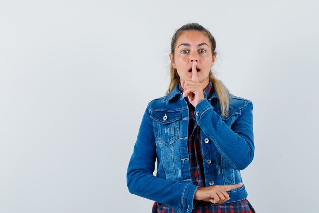 Jeune femme en chemise, veste montrant le geste de silence et l'air confiant, vue de face.